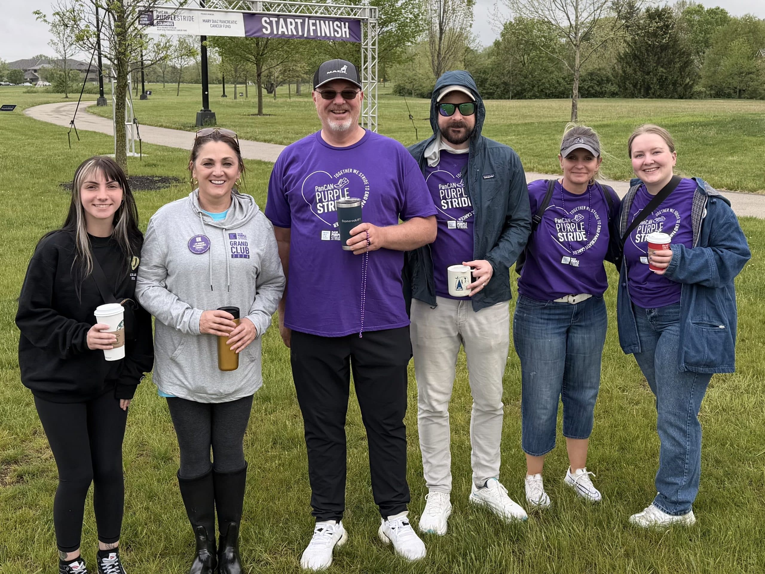 Six people stand together in a grassy park at a Start/Finish area, many wearing purple PanCAN PurpleStride shirts and holding drinks for a charity event. IMG_0022 - Oliver Law Office IMG_0022 - Oliver Law Office