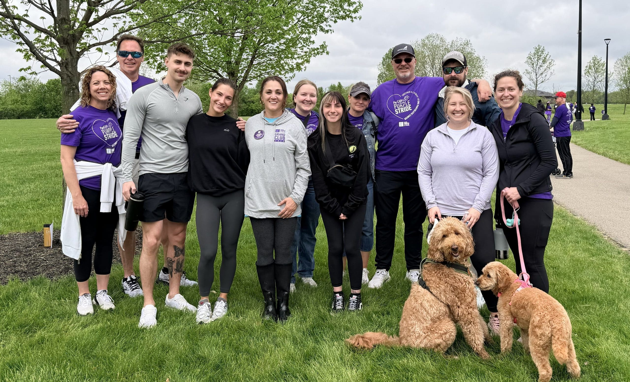 Group of smiling volunteers posing in a park, several wearing purple shirts for Purple Stride; two golden doodles sit at right. IMG_0035 -R - Oliver Law Office IMG_0035 -R - Oliver Law Office