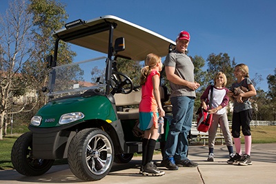 Man in a red cap stands with a group of children near a green golf cart on a sunny day in a park. Lifestyle_Family_Sports - Oliver Law Office