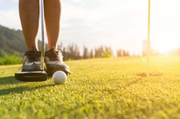 Golfer's feet with a putter ready to strike a golf ball on a sunlit green near the hole. hit-by-golf-ball - Oliver Law Office