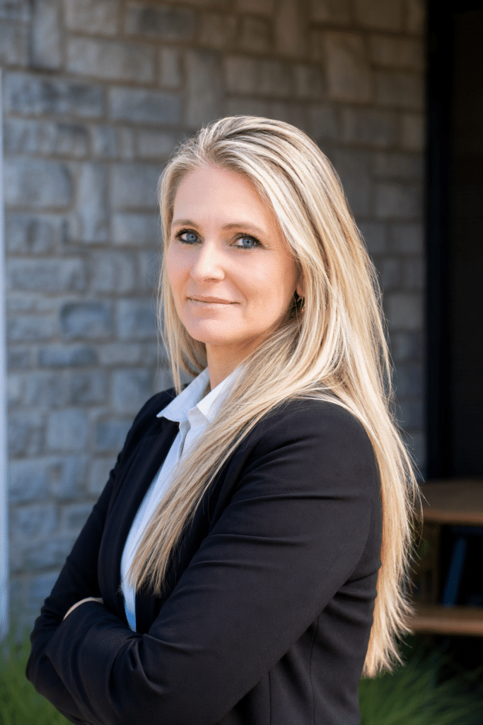 Portrait of a blonde woman in a black blazer and white shirt, looking at the camera outdoors near a stone wall. Angi R photo edit - Oliver Law Office Angi R photo edit - Oliver Law Office