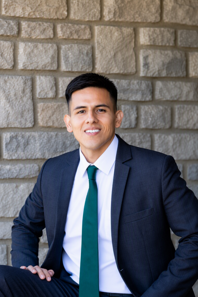 Portrait of a man in a dark suit, white shirt, and green tie, seated against a textured stone wall, smiling at the camera. Juaquin Farid edited - Oliver Law Office Juaquin Farid edited - Oliver Law Office