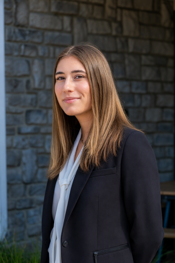 Young woman with long brown hair in a black blazer and white blouse, standing against a stone wall outdoors. Rammel Isabelle Photo edited - Oliver Law Office Rammel Isabelle Photo edited - Oliver Law Office
