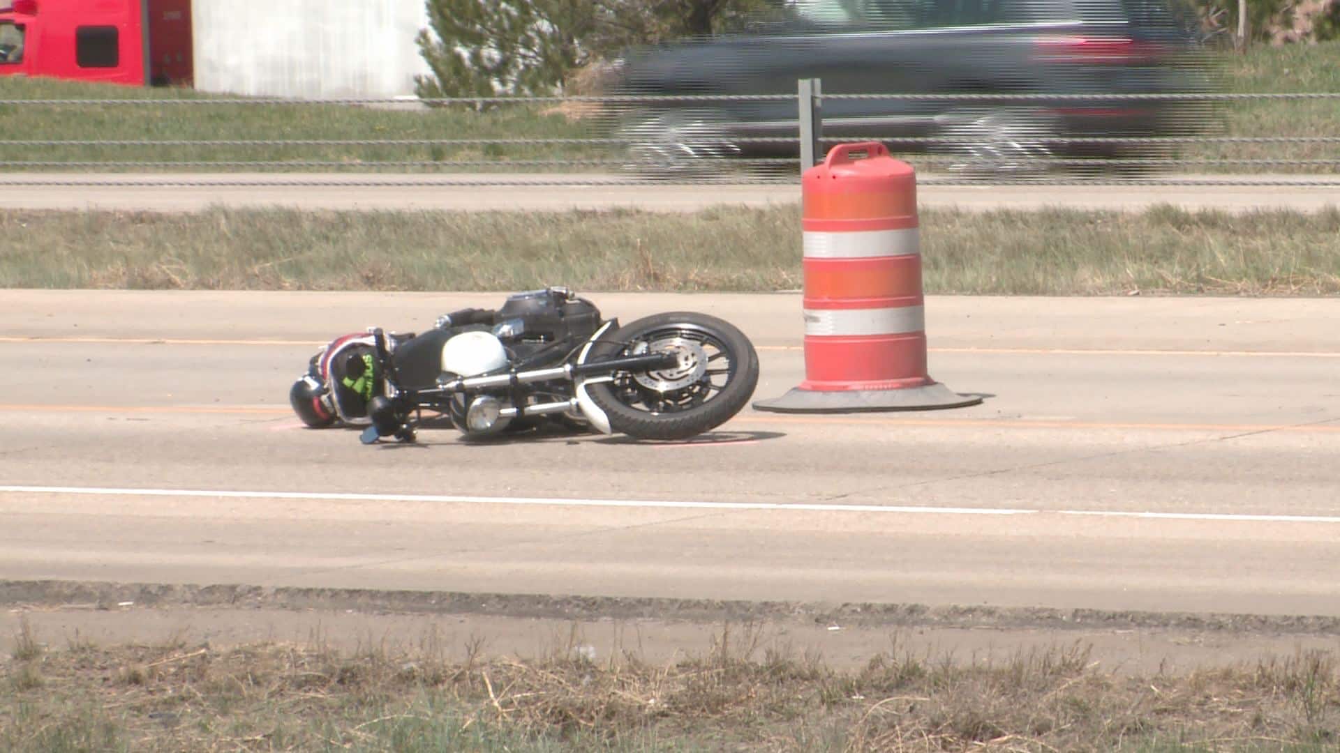 Motorcycle on its side after a crash on a road, with an orange construction barrel nearby and a blurred passing car in the background. motorcycle accident - Oliver Law Office