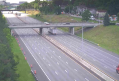 Wide highway with several lanes and an overpass, trucks traveling in both directions, flanked by grassy slopes and trees.