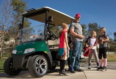 Man in a red cap stands with a group of children near a green golf cart on a sunny day in a park.