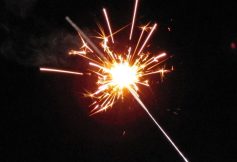 Hand holding a lit sparkler with bright orange-white sparks against a dark background.