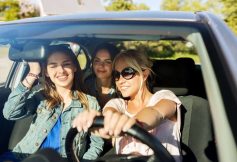 Three friends in a car smiling; the blonde woman in sunglasses drives at the wheel while two others sit in the passenger and back seats on a sunny day.