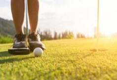 Golfer's feet with a putter ready to strike a golf ball on a sunlit green near the hole.