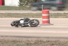 Motorcycle on its side after a crash on a road, with an orange construction barrel nearby and a blurred passing car in the background.